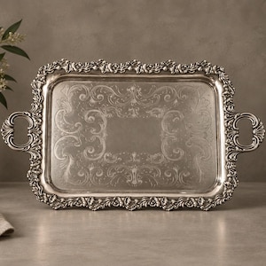 Puede incluir: Una bandeja de servicio rectangular de color plateado con un borde elevado y ornamentado con intrincados detalles florales y de volutas. La bandeja incluye dos asas laterales decorativas y una superficie central grabada con patrones simétricos.