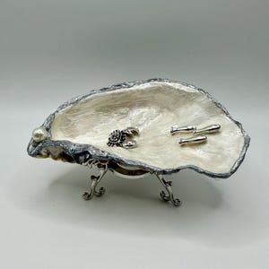 May include: An oyster shell trinket dish with a pearlescent white interior and a silver-painted rim. The dish is supported by a silver-toned metal stand with three curved legs and contains a small silver crab charm and three silver fish charms.