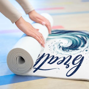 May include: A white yoga mat being rolled up, featuring a blue and teal painted ocean wave graphic and the word "breathe" written in dark blue cursive script. The mat has a textured surface and is shown partially rolled on a light-coloured floor.