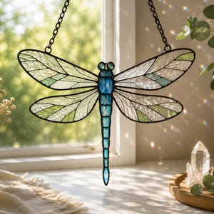 May include: A stained glass dragonfly suncatcher featuring a vibrant blue segmented body and translucent wings with white and light green textured glass panels. The piece is framed in dark metal and hangs from two black chains attached to the top.