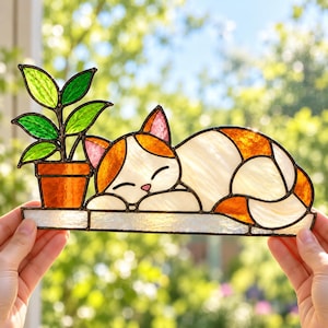 May include: A stained glass suncatcher featuring a sleeping cat with white and orange fur, pink ears, and a small pink nose. Next to the cat is a potted plant with green leaves in an orange pot, all set on a white horizontal base with dark metal lines.