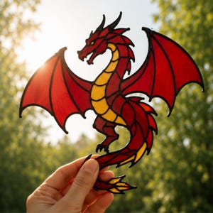 May include: A stained glass suncatcher shaped like a dragon with red wings and a yellow underbelly. The dragon features black lead lines outlining its form, including a long tail, sharp claws, and a detailed head with a pointed snout and horns.