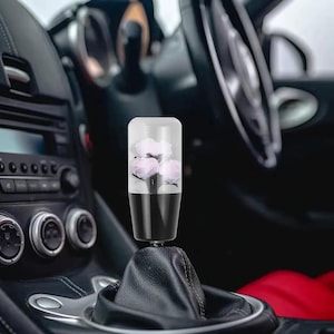May include: A cylindrical car gear shift knob featuring a clear top section that encapsulates three light pink artificial flowers. The base of the knob is solid black and connects to a black leather-like gear shift boot inside a vehicle interior.