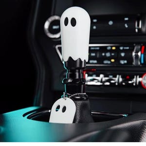 May include: A white ghost-shaped gear shift knob with black oval eyes and a scalloped bottom edge. The knob is mounted on a black metal base featuring the letters WJ and includes a small, matching white ghost-shaped tag on a silver ball chain.