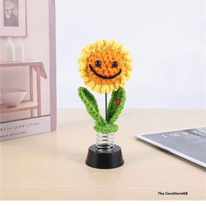 May include: A crocheted sunflower ornament with a yellow petal border, an orange center, and a black stitched smile. The flower features two green leaves, a small red ladybug accent, and is mounted on a silver metal spring atop a black base.