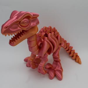 May include: A metallic pink and orange articulated dinosaur figurine with a skeletal structure, featuring a large head with visible teeth, a segmented body, and a long tail. The surface has a shiny, iridescent finish that reflects light.