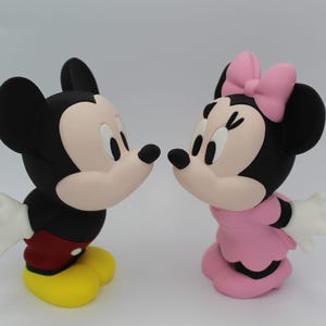 以下が含まれることがあります： 向かい合った2つの漫画キャラクターのフィギュア。左のフィギュアは黒色で、顔はクリーム色、赤いショートパンツ、黄色い靴を履いています。右のフィギュアは黒色で、顔はクリーム色、ピンクのドレスを着て、頭に同色のピンクのリボンをつけています。