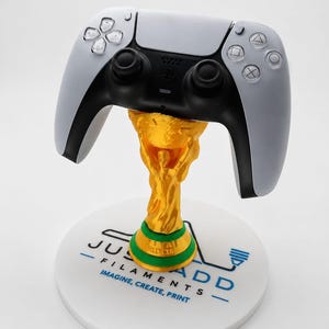 Pode incluir: Um controle de videogame branco e preto apoiado em um suporte de troféu dourado. O suporte possui um anel de base verde e fica sobre uma plataforma circular branca impressa com o texto JUST ADD FILAMENTS IMAGINE, CREATE, PRINT.