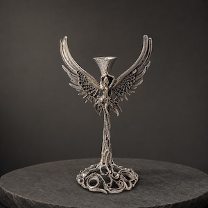 Op de afbeelding: Een zilverkleurige metalen kandelaar met een centrale figuur met uitgestrekte vleugels. De voet lijkt op verstrengelde boomwortels en de bovenkant bevat een getextureerde, conische houder. Het geheel heeft een metallic afwerking met donkere accenten.