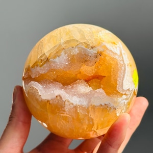 Puede incluir: Una geoda esférica de color amarillo con una superficie exterior pulida y una cavidad central llena de cristales brillantes y translúcidos. El mineral muestra tonos de ámbar dorado y miel, con una pequeña etiqueta amarilla en el lateral.
