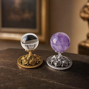 Pode incluir: Dois objetos esféricos em suportes metálicos decorativos. Um é uma esfera de vidro transparente sobre uma base dourada com detalhes em forma de pétalas. O outro é uma esfera de ametista roxa sobre uma base prateada com ornamentos.