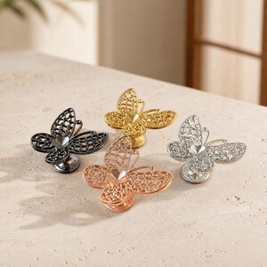 May include: Four butterfly-shaped cabinet knobs in black, gold, rose gold, and silver finishes. Each knob features a delicate, openwork filigree wing design with a small heart-shaped center detail, mounted on a circular base for attachment.