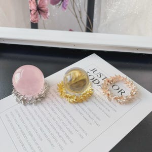 May include: A pink sphere and a clear sphere, each resting on a metallic leaf-shaped stand, alongside a circular metallic leaf-shaped ring. The items are placed on a white card printed with the text JUST TODAY, which lists daily intentions.