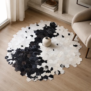 Peut inclure: Un tapis circulaire composé de petites tuiles hexagonales individuelles en peau de vache noire, blanche et grise. Le tapis présente un motif dégradé passant du noir foncé au blanc clair. Un vase en céramique blanc cassé est posé au centre.