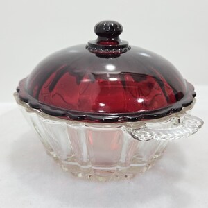 May include: Clear glass candy dish with a ruby red lid and a decorative handle. The lid has a round knob and a scalloped edge. The dish has a clear, faceted bowl.