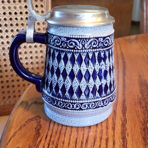 May include: A ceramic beer stein with a blue and white diamond pattern, a blue handle, and a silver metal lid and hinge. The stein has decorative scrollwork around the top and bottom. The stein is sitting on a wooden table.