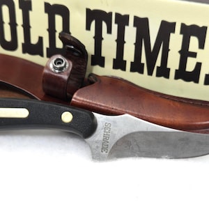 May include: A Schrade Old Timer knife with a black handle and a silver blade, resting in a brown leather sheath. The knife is next to a box with the words "OLD TIMER" in large, bold letters.