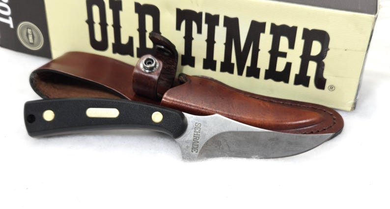 May include: A Schrade Old Timer knife with a black handle and a silver blade, resting in a brown leather sheath. The knife is next to a box with the words "OLD TIMER" in large, bold letters.