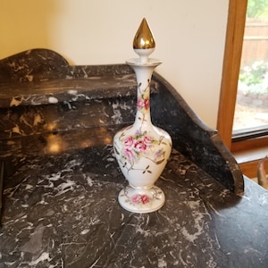 May include: A white porcelain perfume bottle with a gold-colored stopper. The bottle is decorated with pink roses and green leaves. The bottle is approximately 8 inches tall.