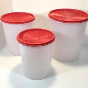 May include: Three cylindrical storage containers with translucent white bodies and circular red lids. Each lid features a textured, sunburst-like pattern radiating from the center. The containers are arranged in a group of three, varying in size.