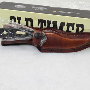 May include: A black-handled knife in a brown leather sheath, with a black button clasp. The knife is partially visible, wrapped in plastic. The sheath is stitched along the edges. The box behind the knife reads "Old Timer" in large black letters.