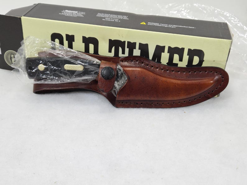 May include: A black-handled knife in a brown leather sheath, with a black button clasp. The knife is partially visible, wrapped in plastic. The sheath is stitched along the edges. The box behind the knife reads "Old Timer" in large black letters.