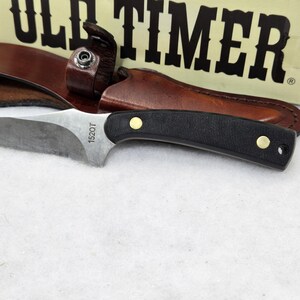 May include: A hunting knife with a black handle and a silver blade, resting on a white surface. The knife is accompanied by a brown leather sheath. The text "1520T" is visible on the blade. The box behind the knife reads "OLD TIMER".