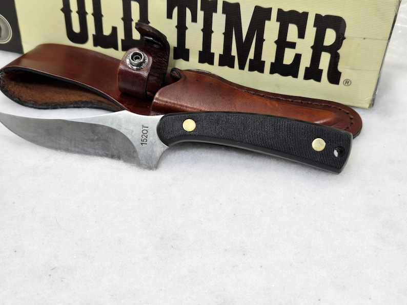 May include: A hunting knife with a black handle and a silver blade, resting on a white surface. The knife is accompanied by a brown leather sheath. The text "1520T" is visible on the blade. The box behind the knife reads "OLD TIMER".