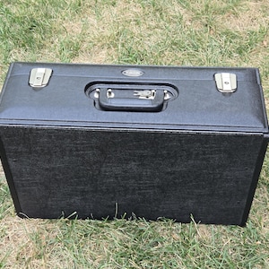 May include: A rectangular black hard-shell carrying case with a textured finish. The case features a black plastic handle, a central locking mechanism with a small silver key inserted, and two silver metal latches on the top lid surface.