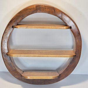 May include: A round, wooden wall shelf with three shelves. The frame is a dark brown, while the shelves are a lighter wood tone. The shelf is designed with a circular shape, adding a unique touch to any room. The wood grain is visible, giving it a rustic feel.