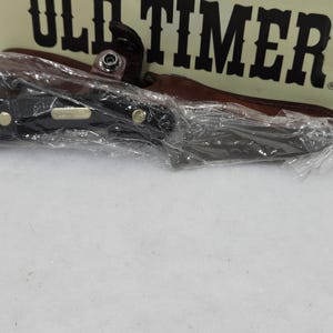 May include: A black folding knife with a brown leather sheath, packaged in clear plastic. The knife has a black handle with gold-colored rivets. The packaging includes the text "OLD TIMER".