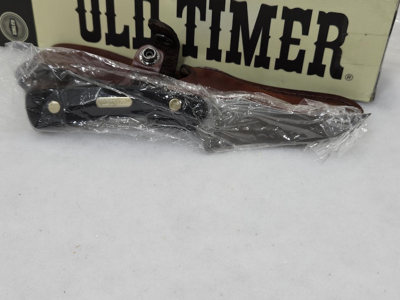May include: A black folding knife with a brown leather sheath, packaged in clear plastic. The knife has a black handle with gold-colored rivets. The packaging includes the text "OLD TIMER".