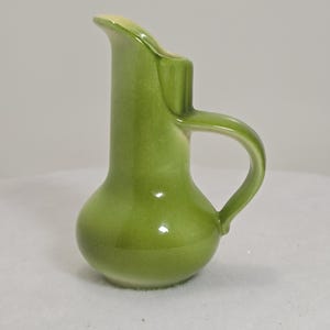 May include: A small, green ceramic pitcher with a rounded base and a curved handle. The pitcher has a spout for pouring and a glossy finish. The color is a vibrant, spring-like green, with a hint of yellow at the spout.