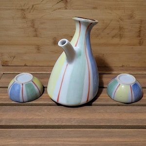 May include: A ceramic sake set featuring a tall, slender pitcher with a spout and two small matching cups. The set is decorated with vertical stripes in pastel colors, including blue, yellow, green, and red. The pitcher has a brown rim.