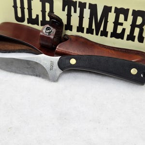 May include: A fixed-blade knife with a black handle and a silver blade, accompanied by a brown leather sheath. The blade is marked with "1520T". The knife is set against a backdrop featuring the "OLD TIMER" logo.