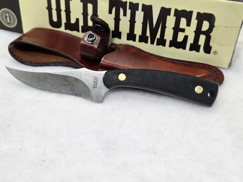 May include: A fixed-blade knife with a black handle and a silver blade, accompanied by a brown leather sheath. The blade is marked with "1520T". The knife is set against a backdrop featuring the "OLD TIMER" logo.