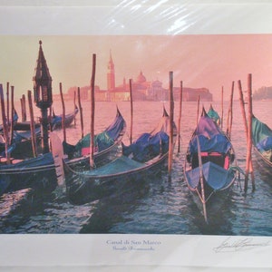 May include: A print of the Canal di San Marco in Venice, Italy, featuring gondolas docked in the water. The image has a warm color palette with a pink and orange sky. The gondolas have blue and green covers. The print is signed.