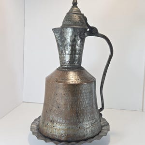 May include: A tall, hammered metal pitcher with a wide, cylindrical copper-toned base and a narrower, silver-toned neck. It features a hinged lid with a pointed finial, a curved handle, and sits on a circular, scalloped-edge metal tray base.