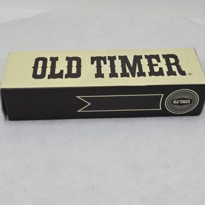 May include: Rectangular box with the words "OLD TIMER" in bold, dark brown letters on a cream-colored background. The box has a black band with a decorative arrow and a circular emblem that reads "OLD TIMER".