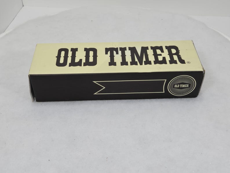 May include: Rectangular box with the words "OLD TIMER" in bold, dark brown letters on a cream-colored background. The box has a black band with a decorative arrow and a circular emblem that reads "OLD TIMER".