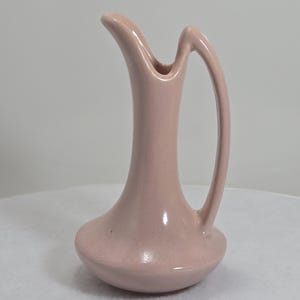 May include: A small, pink ceramic vase with a unique, curved handle and spout. The vase has a bulbous base and a slender neck, with a smooth, glossy finish. The overall design is minimalist and elegant, suitable for displaying flowers or as a decorative piece.