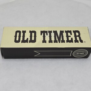 May include: A rectangular box with the words "OLD TIMER" in bold, dark brown letters. The top of the box is a light tan color, and the bottom is black. The box has a banner design and the words "OLD TIMER" in a circle.
