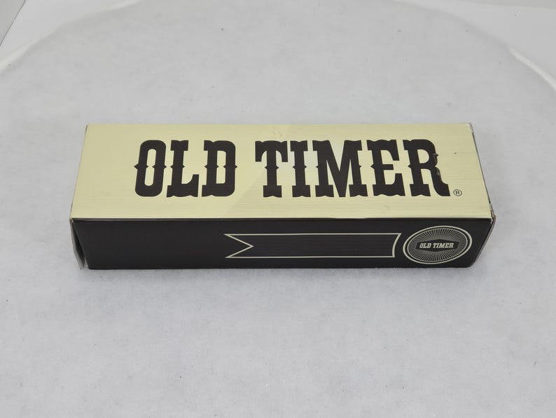 May include: A rectangular box with the words "OLD TIMER" in bold, dark brown letters. The top of the box is a light tan color, and the bottom is black. The box has a banner design and the words "OLD TIMER" in a circle.