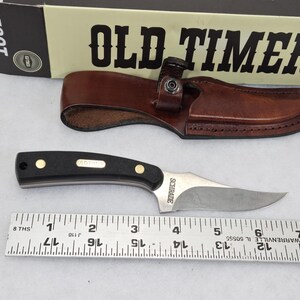 May include: A black and silver knife with a brown leather sheath, and a box labeled "OLD TIMER". The knife has a black handle with gold accents and a silver blade. A ruler is below the knife.