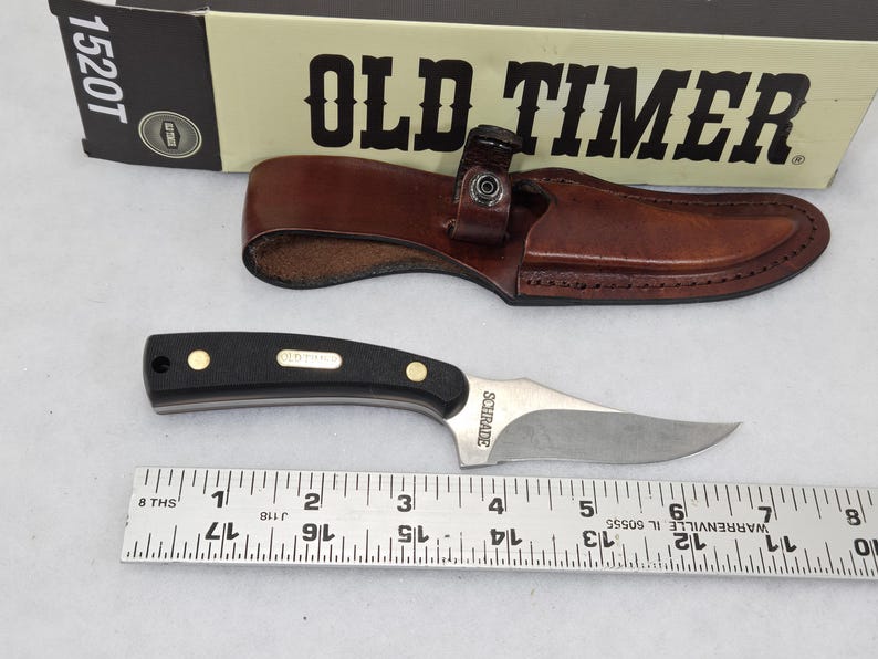 May include: A black and silver knife with a brown leather sheath, and a box labeled "OLD TIMER". The knife has a black handle with gold accents and a silver blade. A ruler is below the knife.