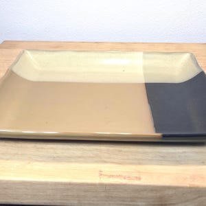 May include: Rectangular ceramic serving platter with a color-block design. The platter is divided into three sections: beige, tan, and dark gray. The edges are slightly curved, and the surface is glossy.