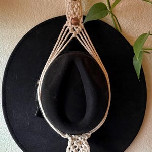 May include: A black wide-brimmed hat held by a cream-colored macrame hanger. The hanger features a wooden ring at the top, three brown wooden beads integrated into the knotted cord design, and a fringe finish at the bottom of the structure.