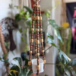 May include: A hanging plant holder constructed from strands of wooden beads in natural, dark brown, and patterned red tones. The beads are strung on twine and suspended from a wooden ring, supporting a small, square white ceramic planter at the base.