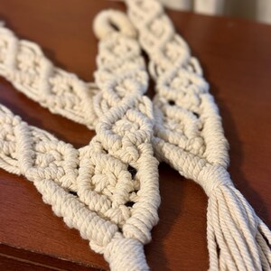 May include: Three off-white macrame straps crafted from thick cotton cord, featuring intricate knotted diamond patterns and finished with long, loose fringe tassels at the bottom ends. Each strap includes a small loop at the top for hanging.