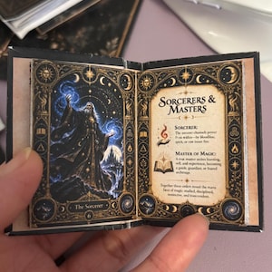 May include: A miniature open book featuring an illustration of a sorcerer casting blue magic on the left page and the text Sorcerers & Masters with descriptions of a sorcerer and master of magic on the right page, framed by gold celestial motifs.